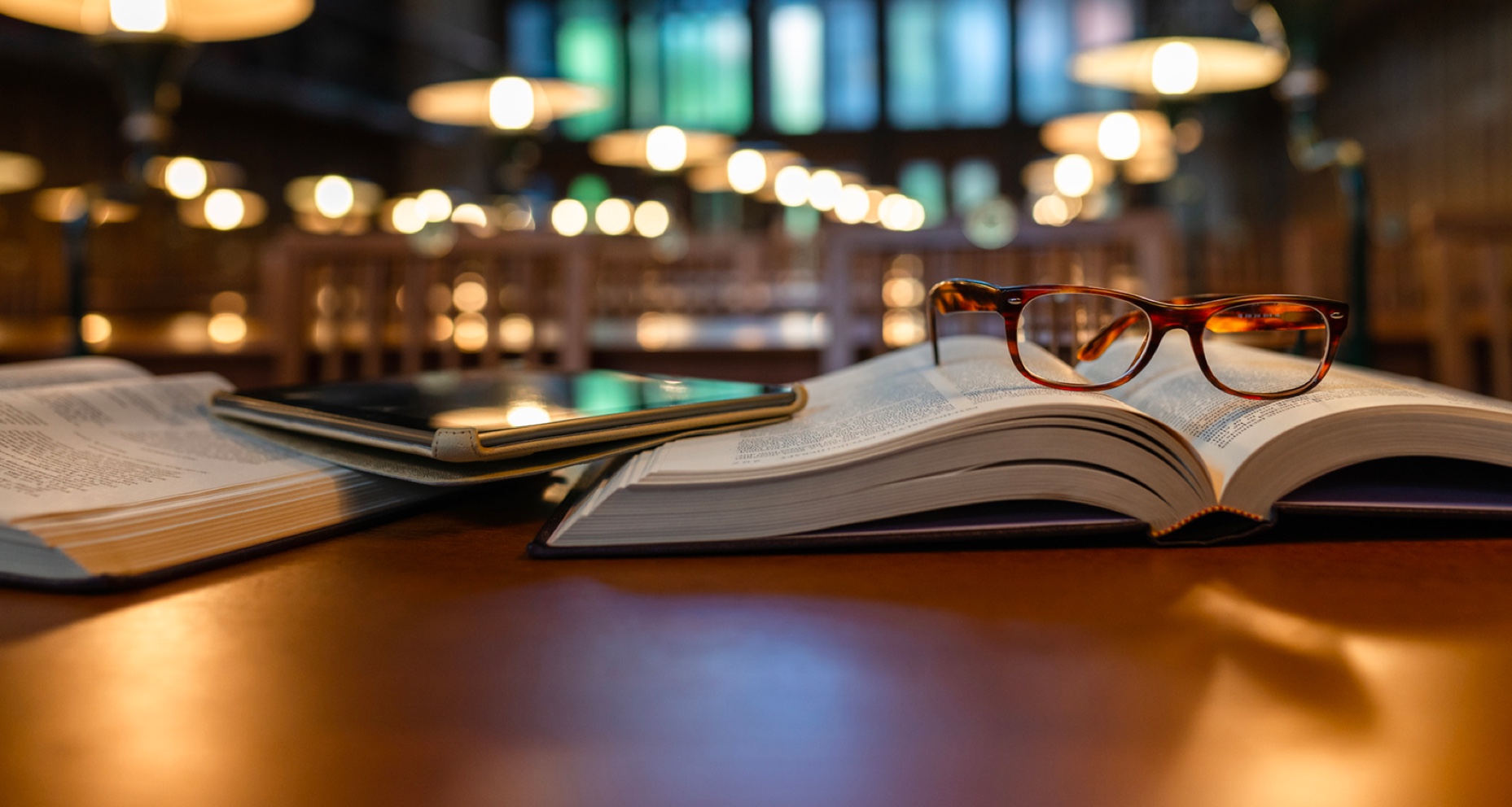 A pair of glasses on top of a book and e-tablet on a desk in a library.
